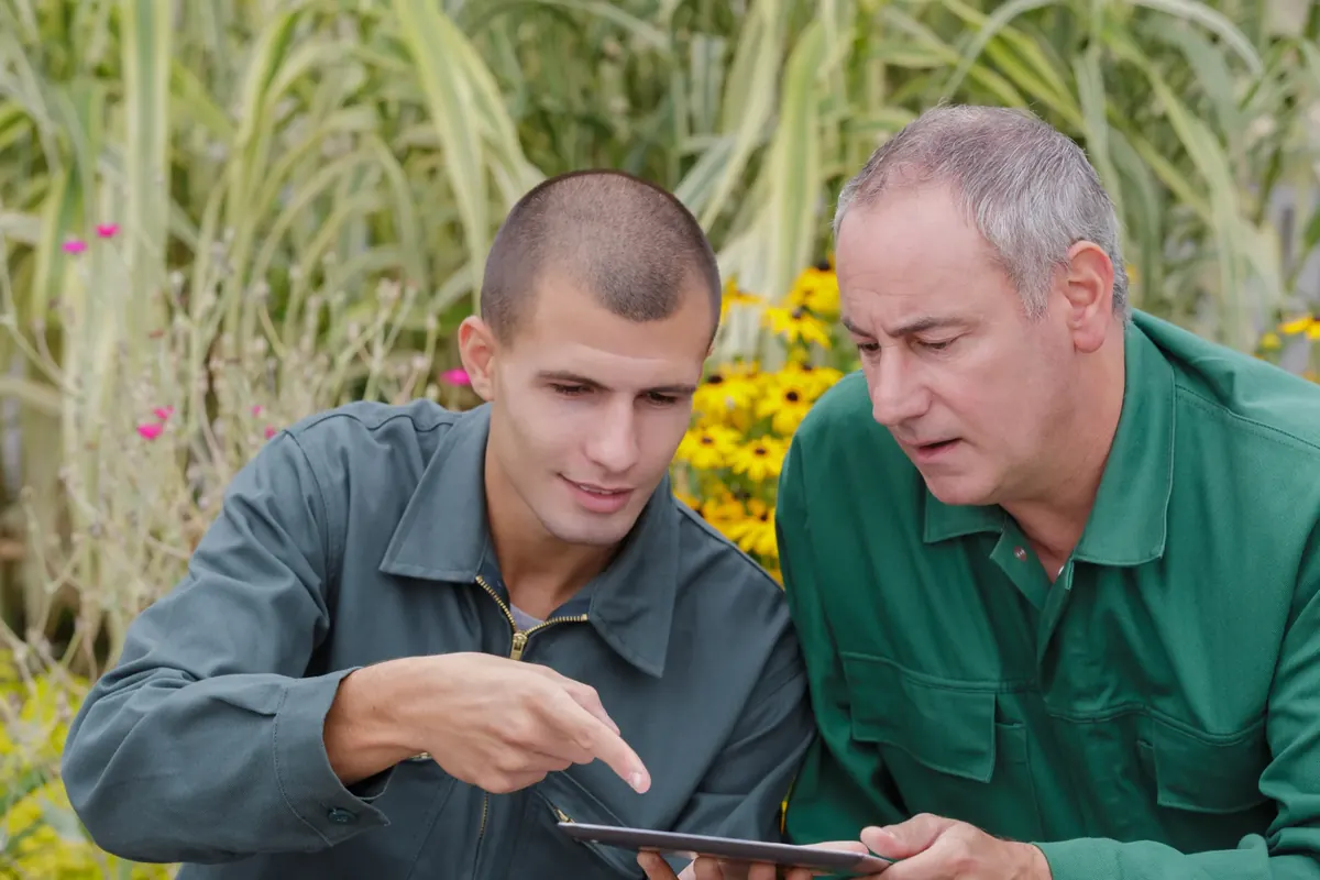 Two people in work jackets leaning in together over a tablet, going through it line by line, garden planting behind them.